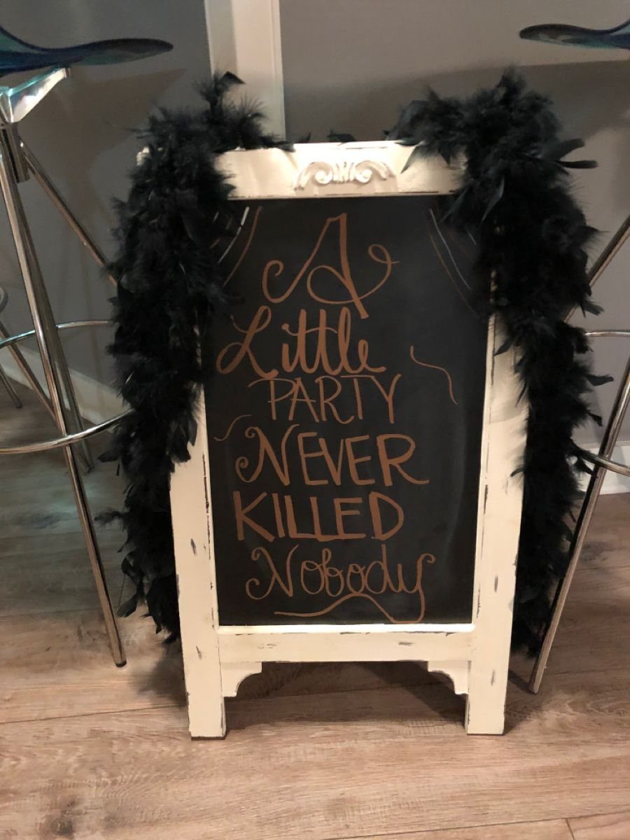 A chalkboard sign with gold lettering framed by black feather boas on a wooden floor.