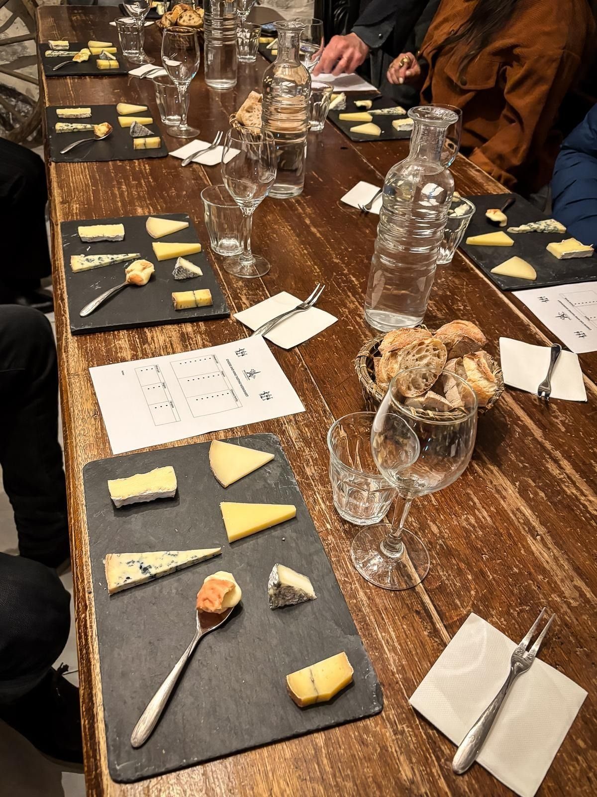 Various cheese slices on slate platters arranged on a long wooden table with bread and wine glasses.