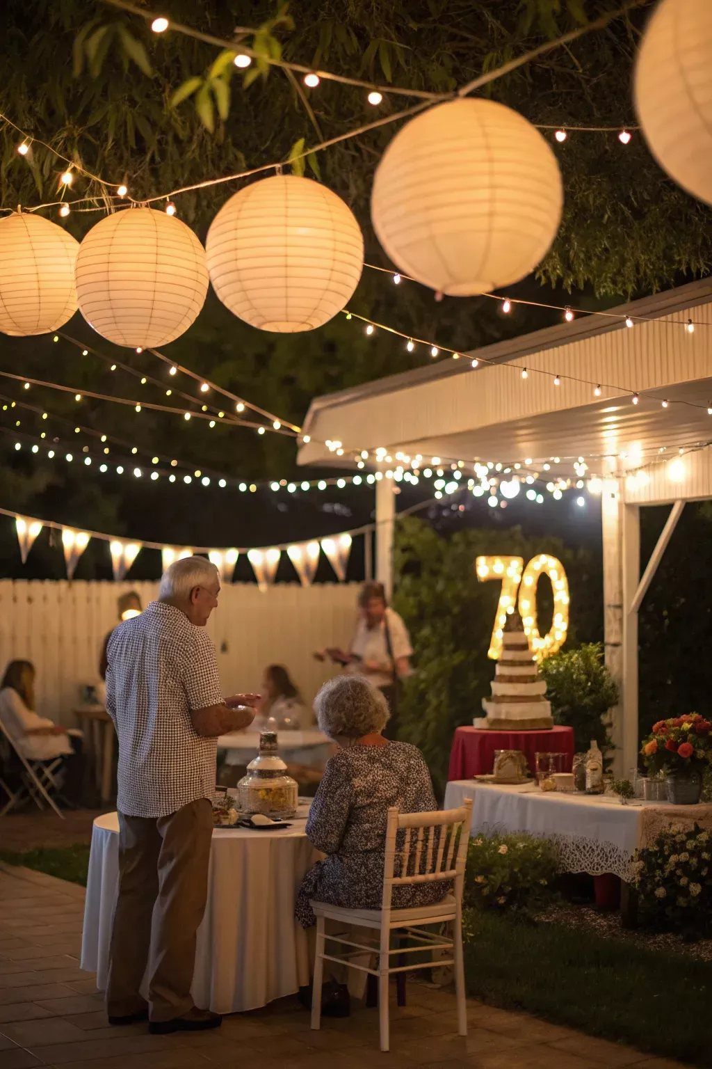 Large white paper lanterns and string lights hanging over an outdoor evening birthday celebration.