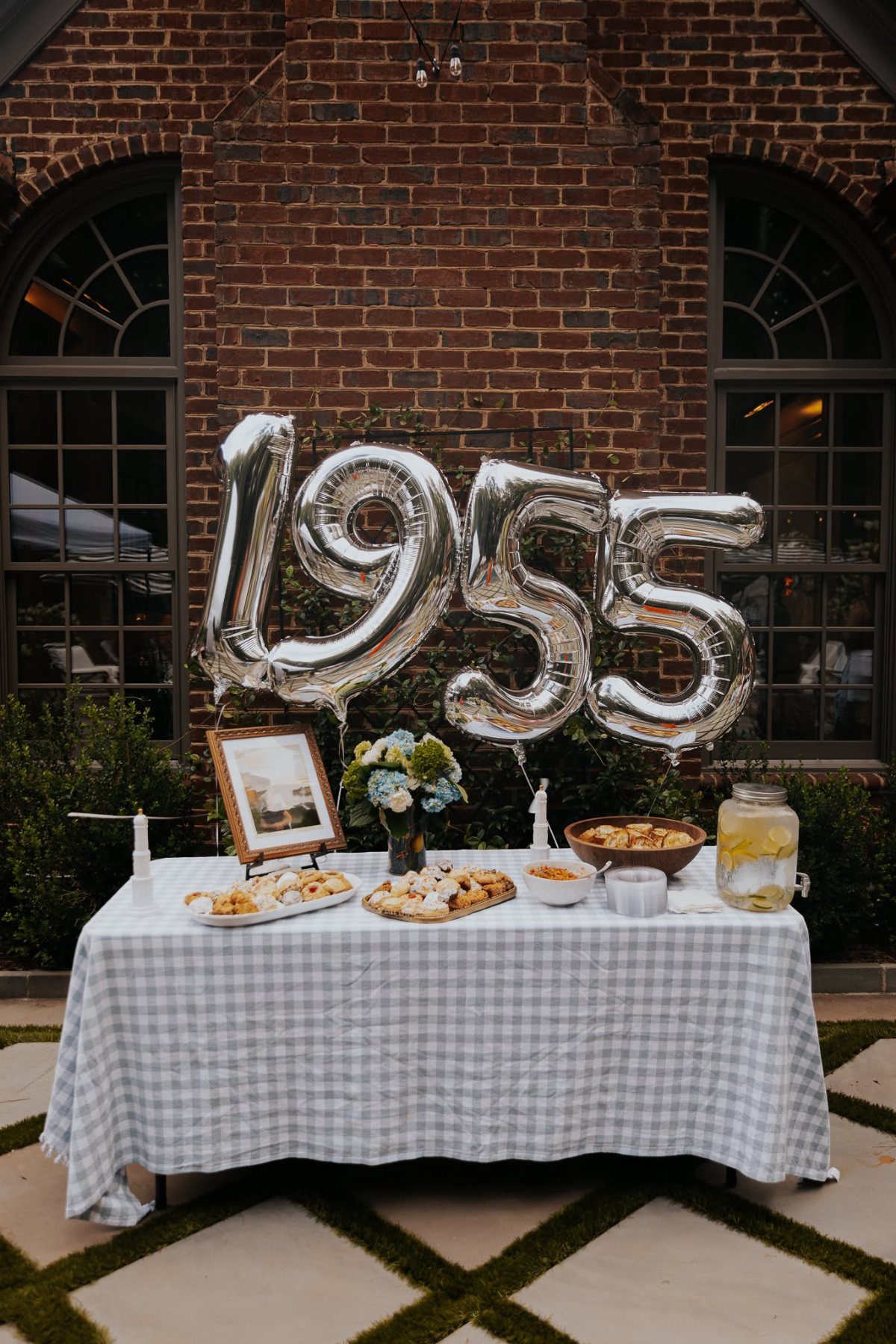 Silver balloons spelling 1955 stand behind a snack table with a checkered tablecloth outdoors.