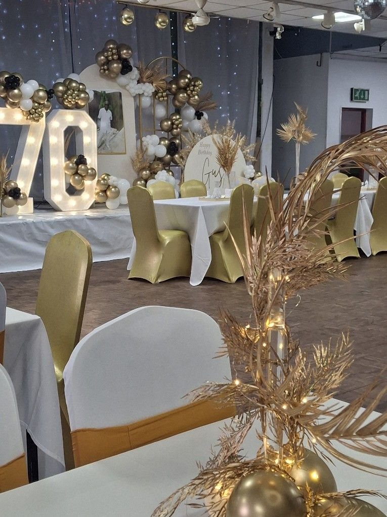 Large illuminated numbers seventy surrounded by gold and white balloons and decorated banquet tables.