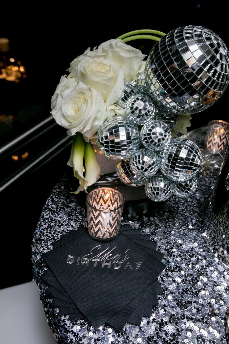 White roses and silver disco balls arranged on a sequined tablecloth with black napkins.