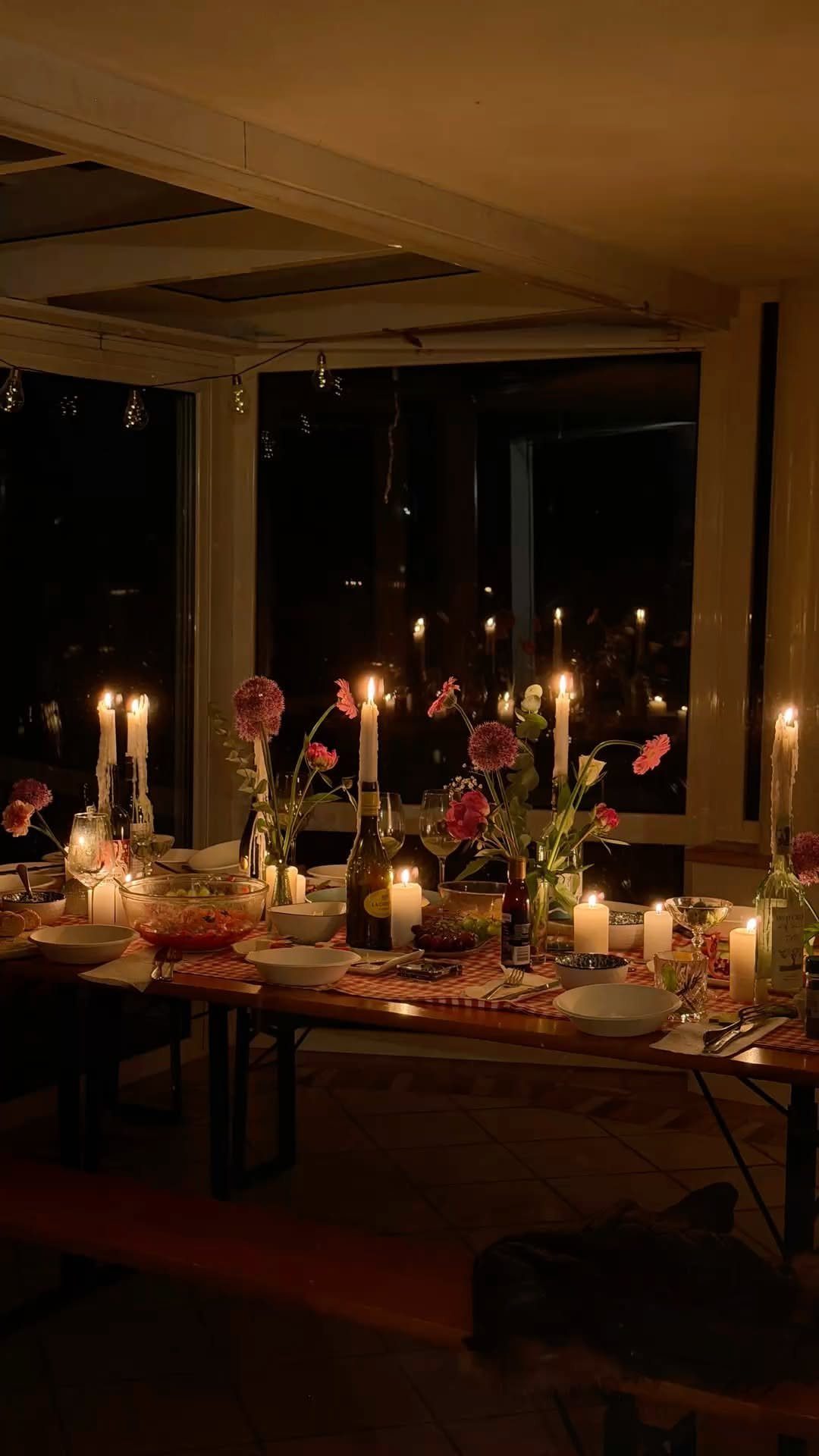 A long wooden dining table set with lit candles, flowers, and wine for an intimate dinner.