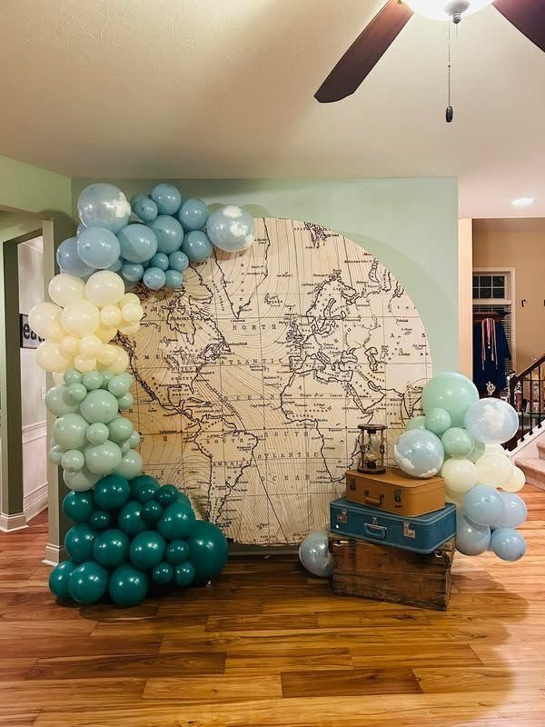A large world map backdrop framed by blue and teal balloon arches and vintage suitcases.