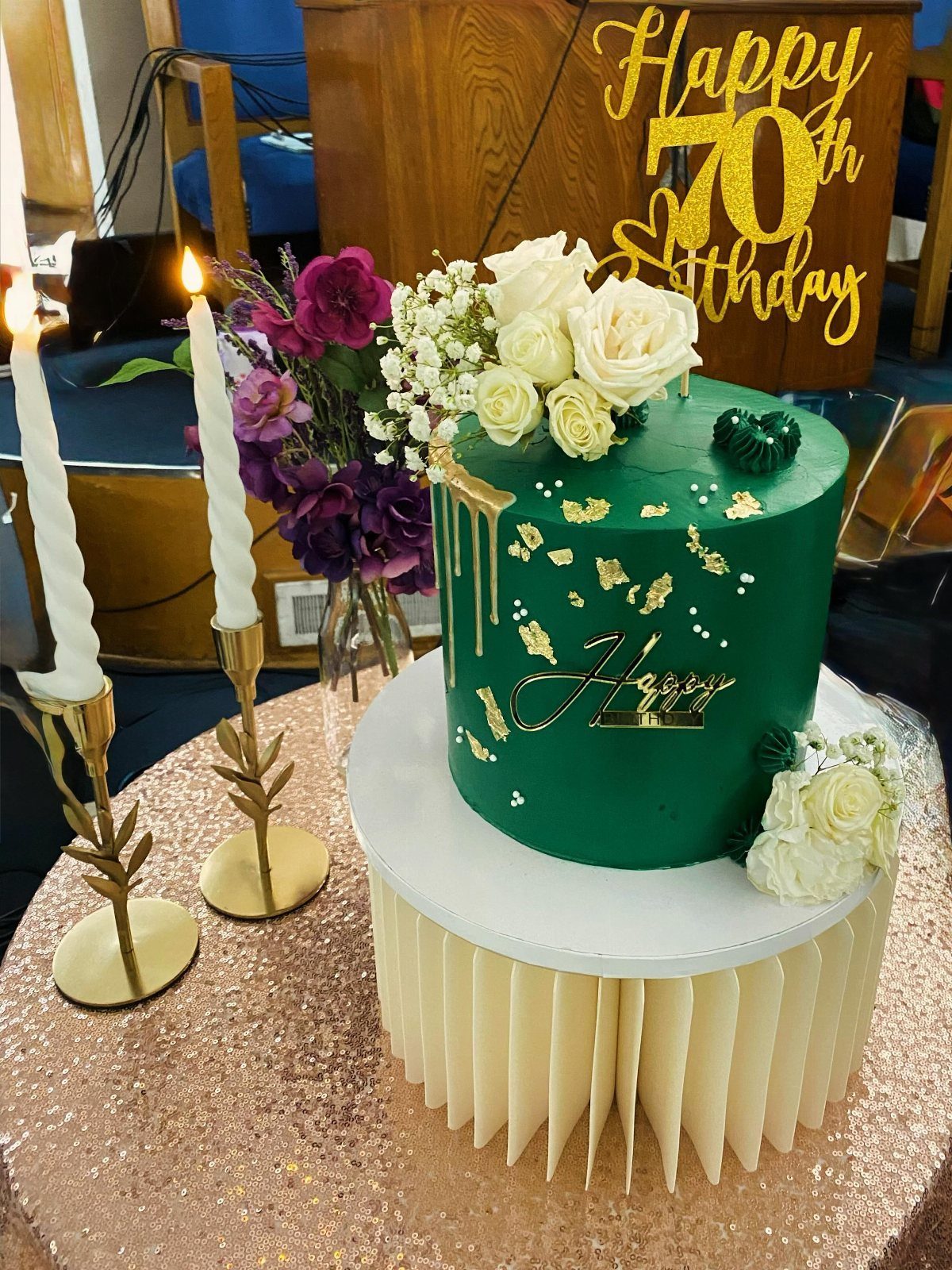 Green 70th birthday cake with gold leaf and topper on rose gold sequin table with candles and flowers.