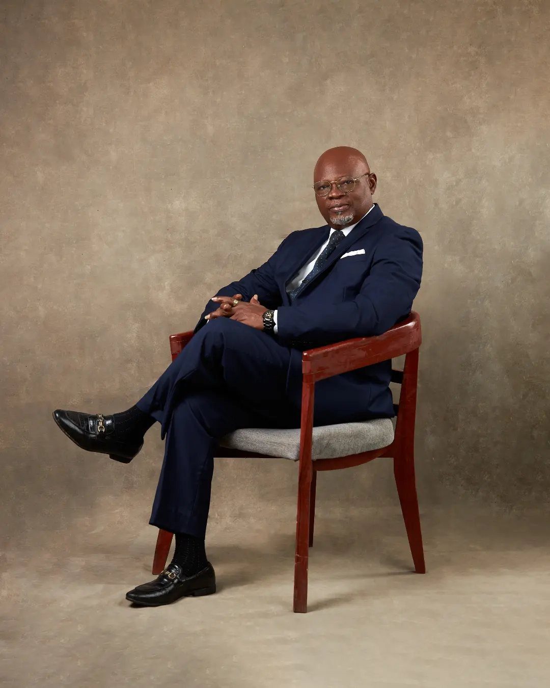 An older man in a navy blue suit sitting in a wooden chair against a brown background.