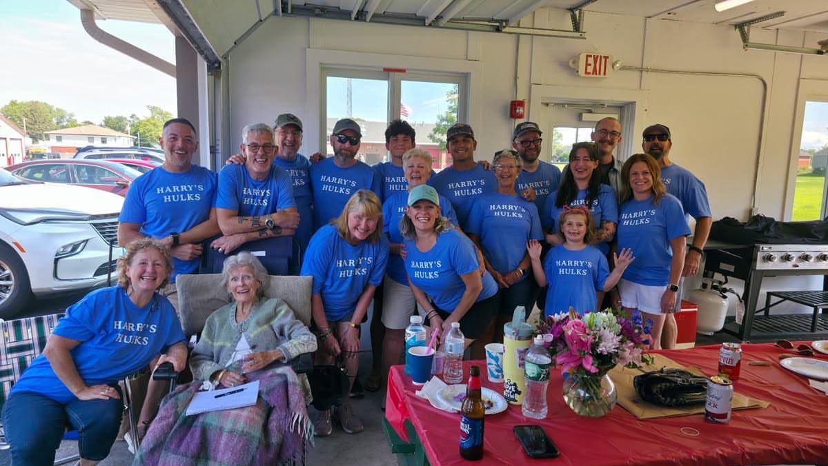 Large group of people in matching blue shirts posing in a garage with an elderly woman seated in front near a party table.