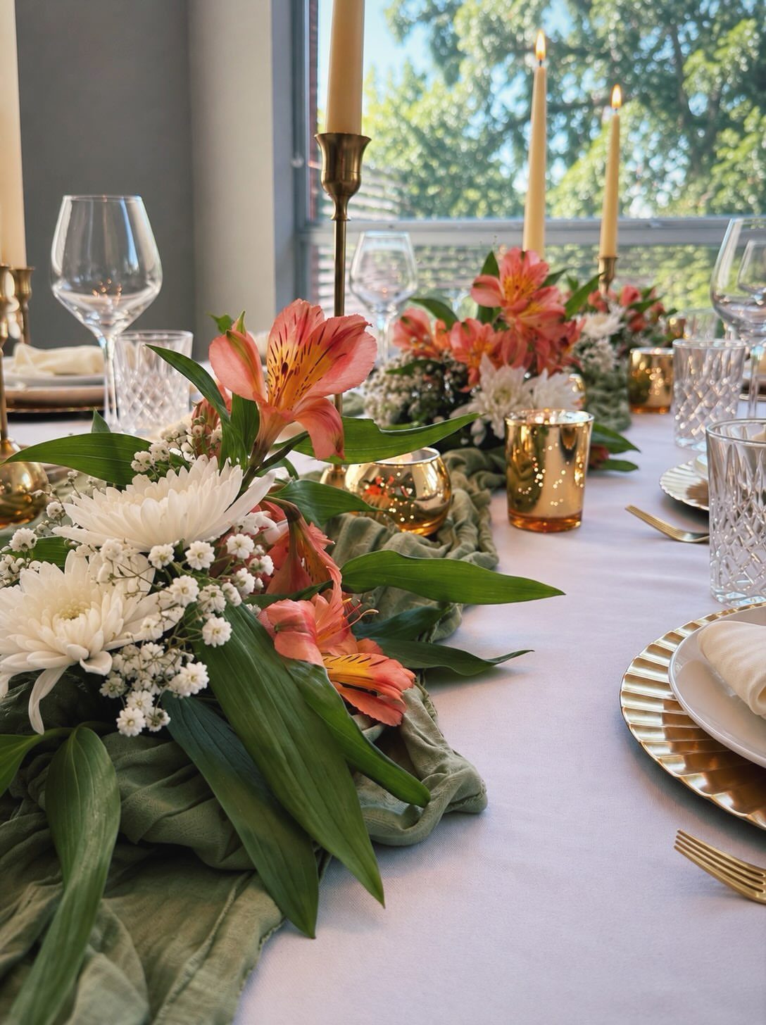 Elegant dinner table setting with pink flowers, gold candles, crystal glassware, and gold-rimmed plates on a white tablecloth.