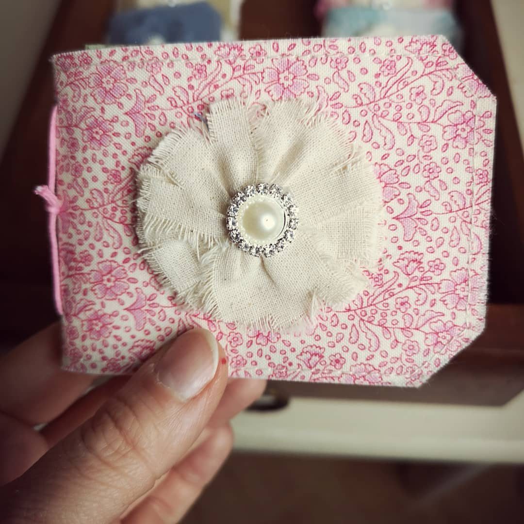 Hand holding small handmade fabric book with pink floral cover and cream fabric flower embellishment featuring pearl center.