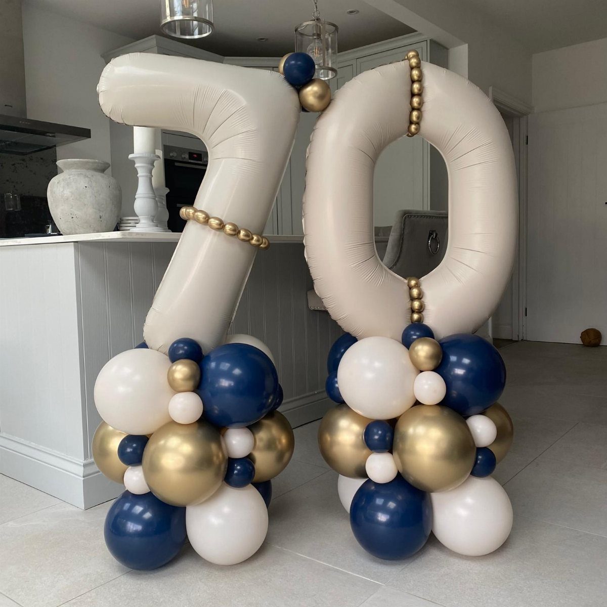 Large cream foil 70 balloons decorated with navy, gold, and white balloon clusters standing in a modern kitchen.