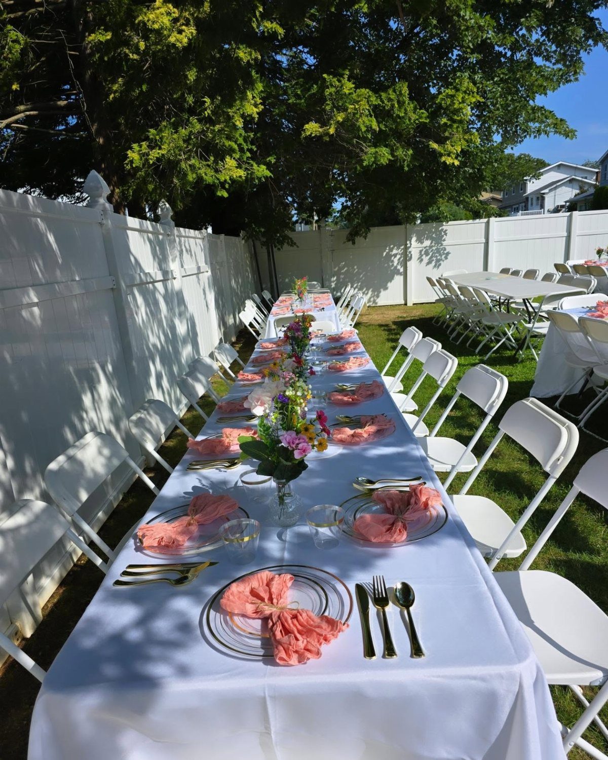 A long outdoor dining table set with white linens, pink napkins, and flowers on a lawn.