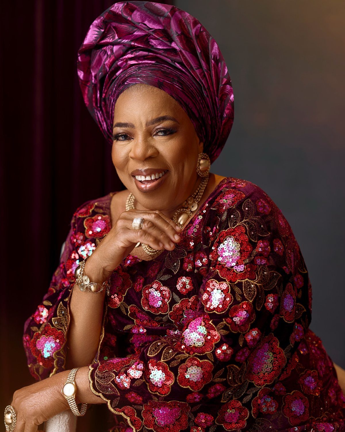 Smiling woman in ornate purple traditional African attire and matching headwrap posing for a portrait.