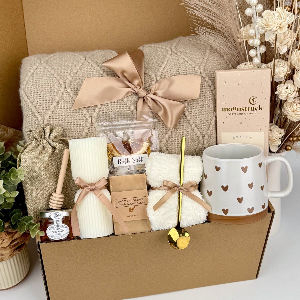 A gift box containing a textured blanket, heart-patterned mug, rolled towels, and bath salts.