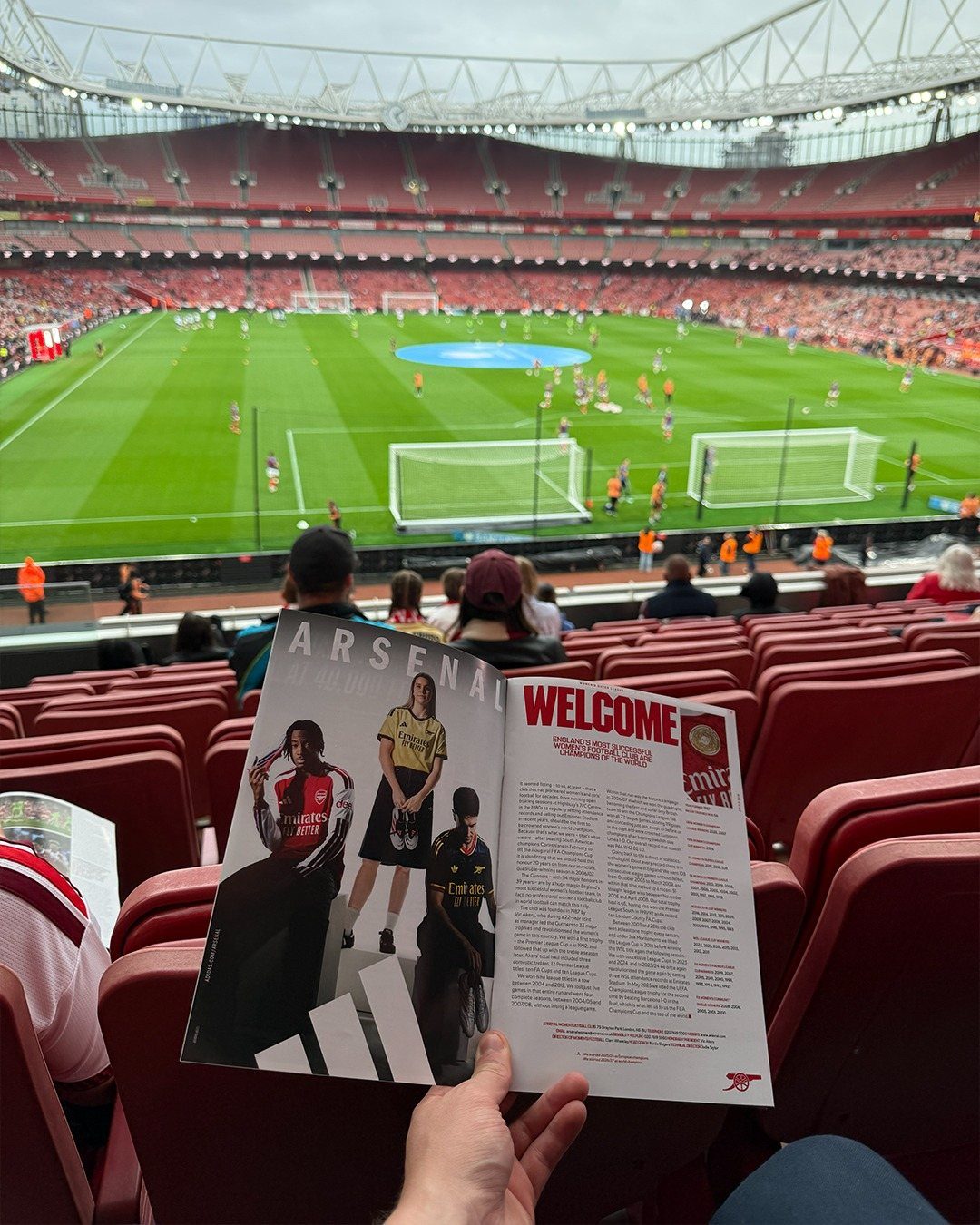 An open Arsenal football program held in front of a large, crowded green football stadium.