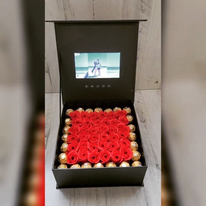 An open black gift box filled with red roses and gold chocolates with a video screen.