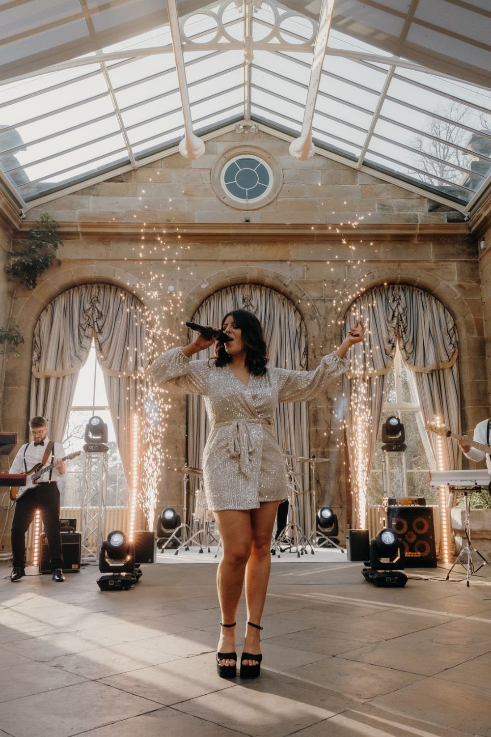 A female singer performing with a microphone amidst sparkling pyrotechnics in a bright conservatory.