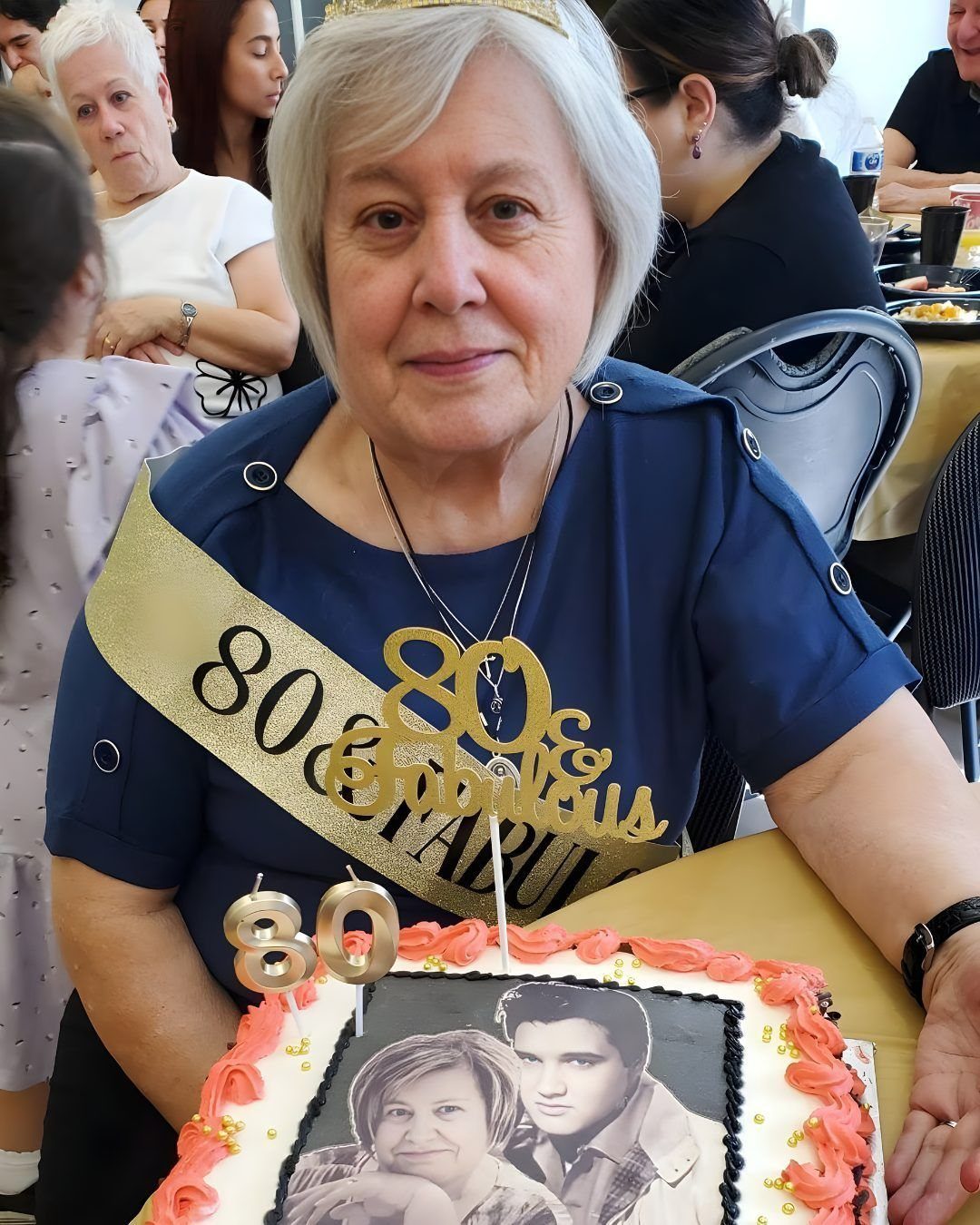 An elderly woman wearing an 80th birthday sash sits behind a decorated birthday cake.