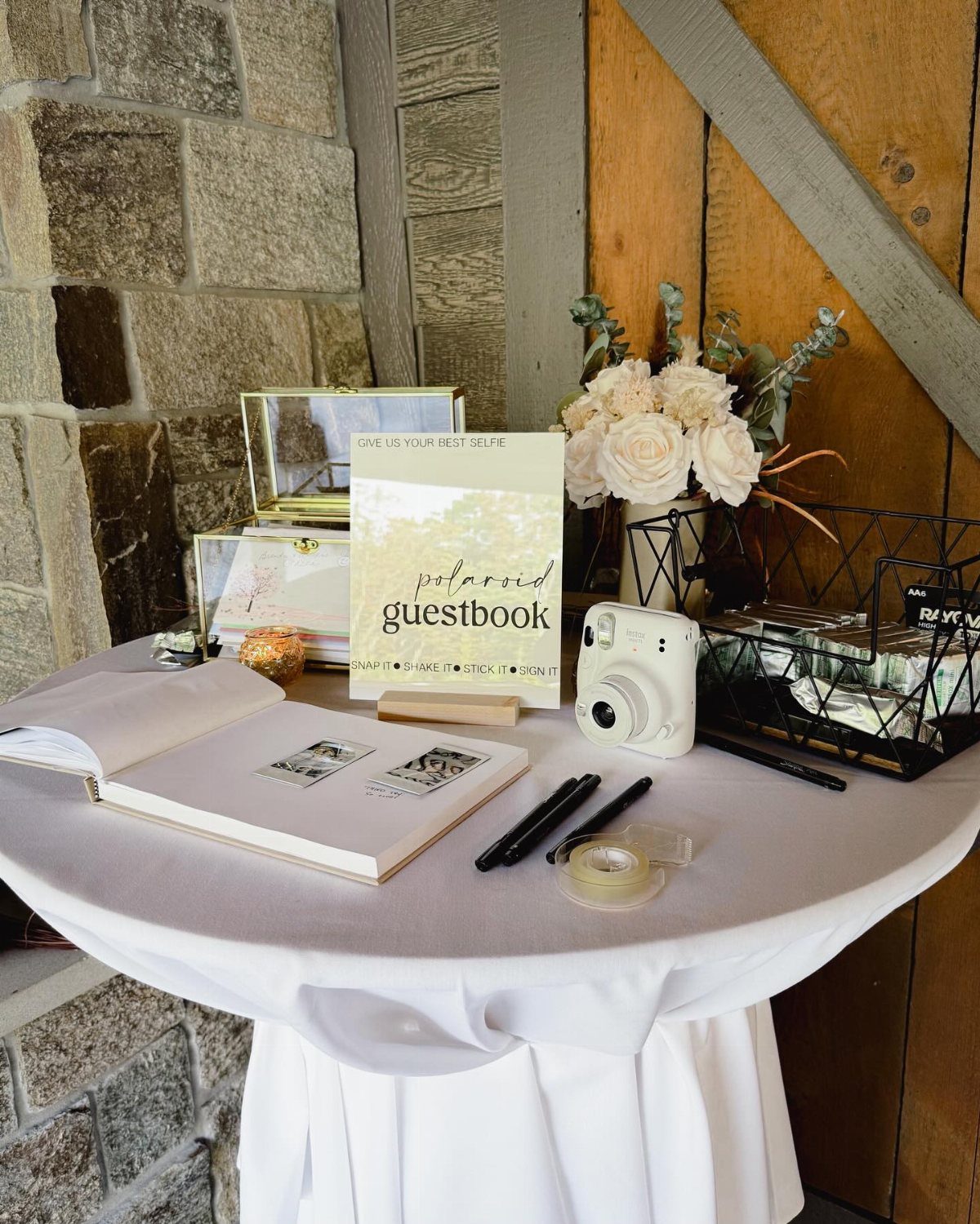 A white guestbook station with an instant camera, markers, and white roses on a draped table.