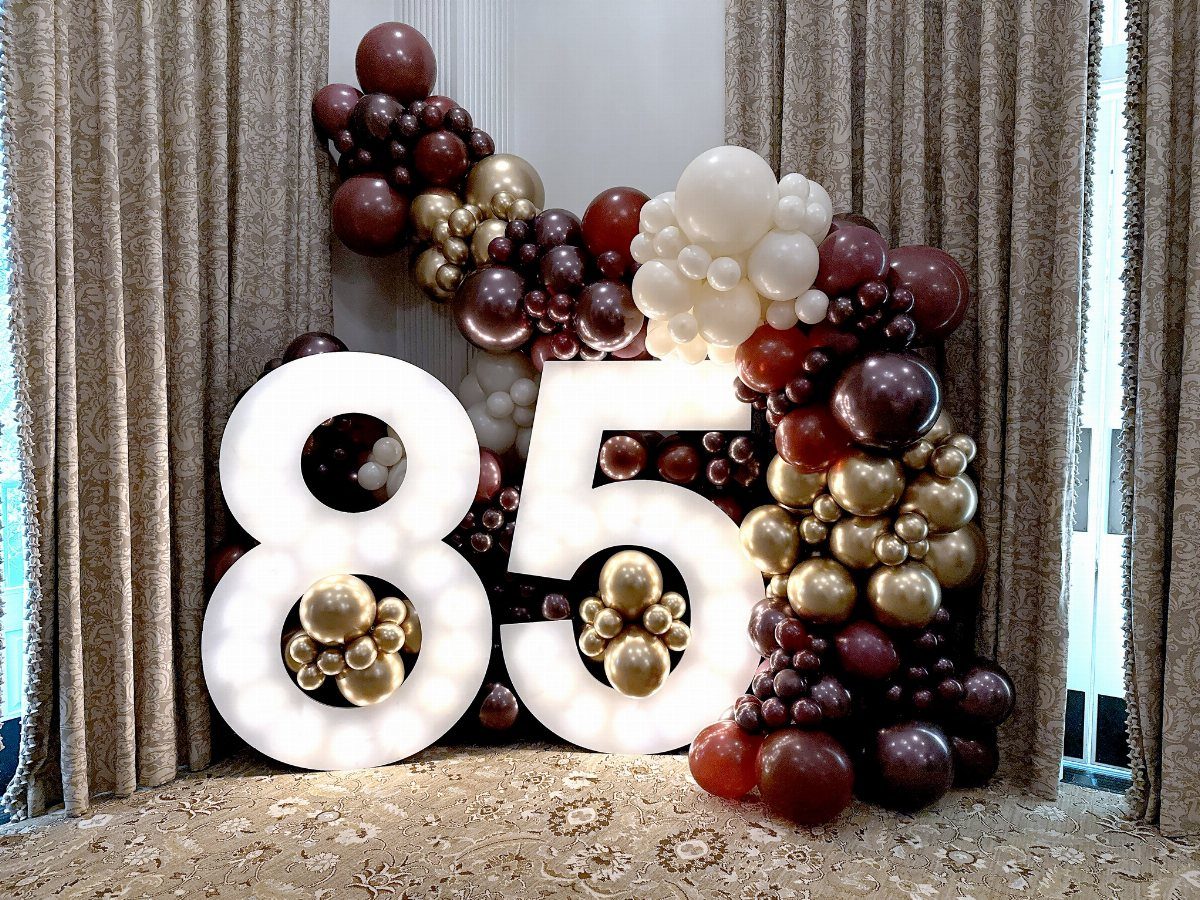 Large illuminated numbers 85 surrounded by burgundy, gold, and white balloons in a decorated room.
