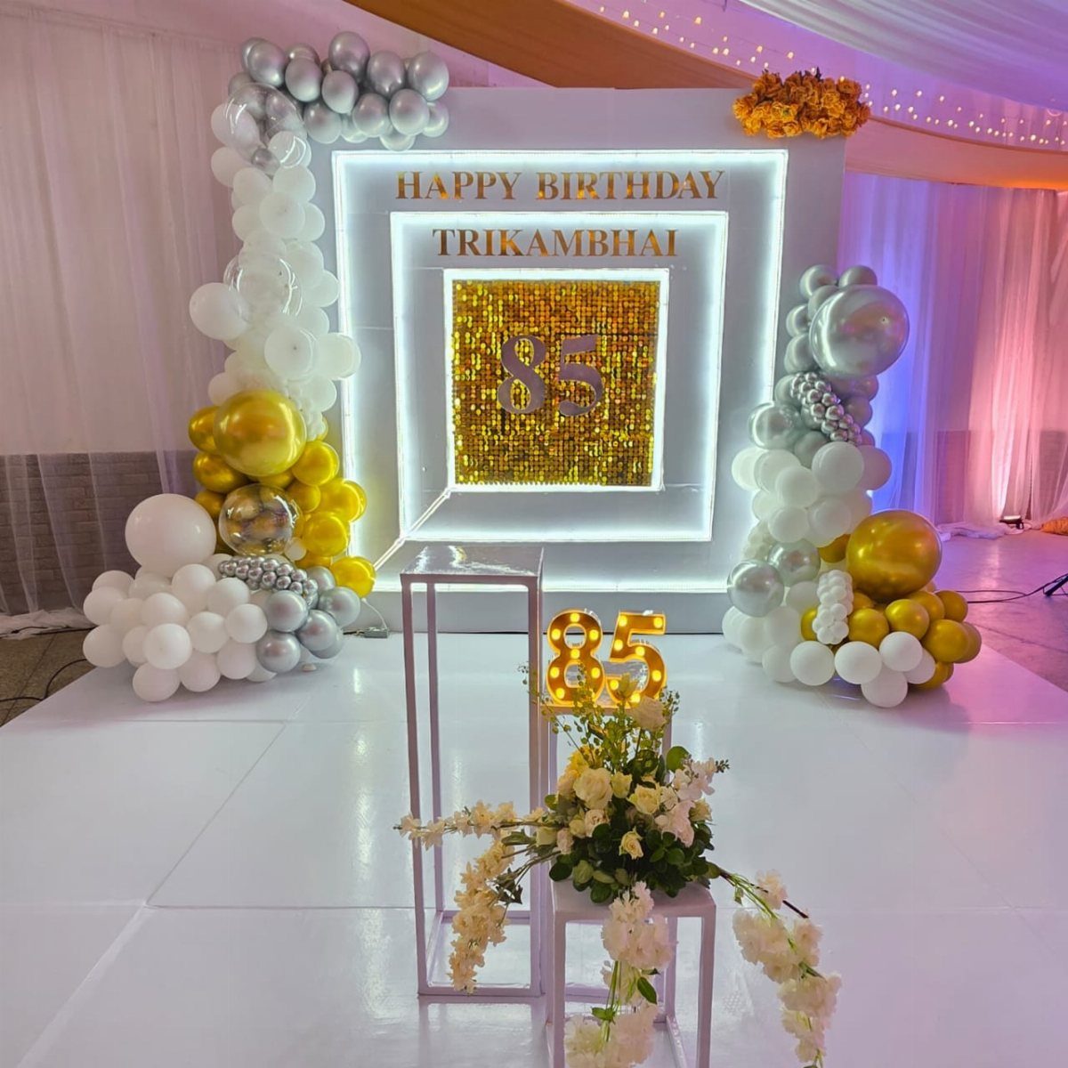 White and gold balloon decorations surrounding an illuminated birthday backdrop with the number eighty-five.