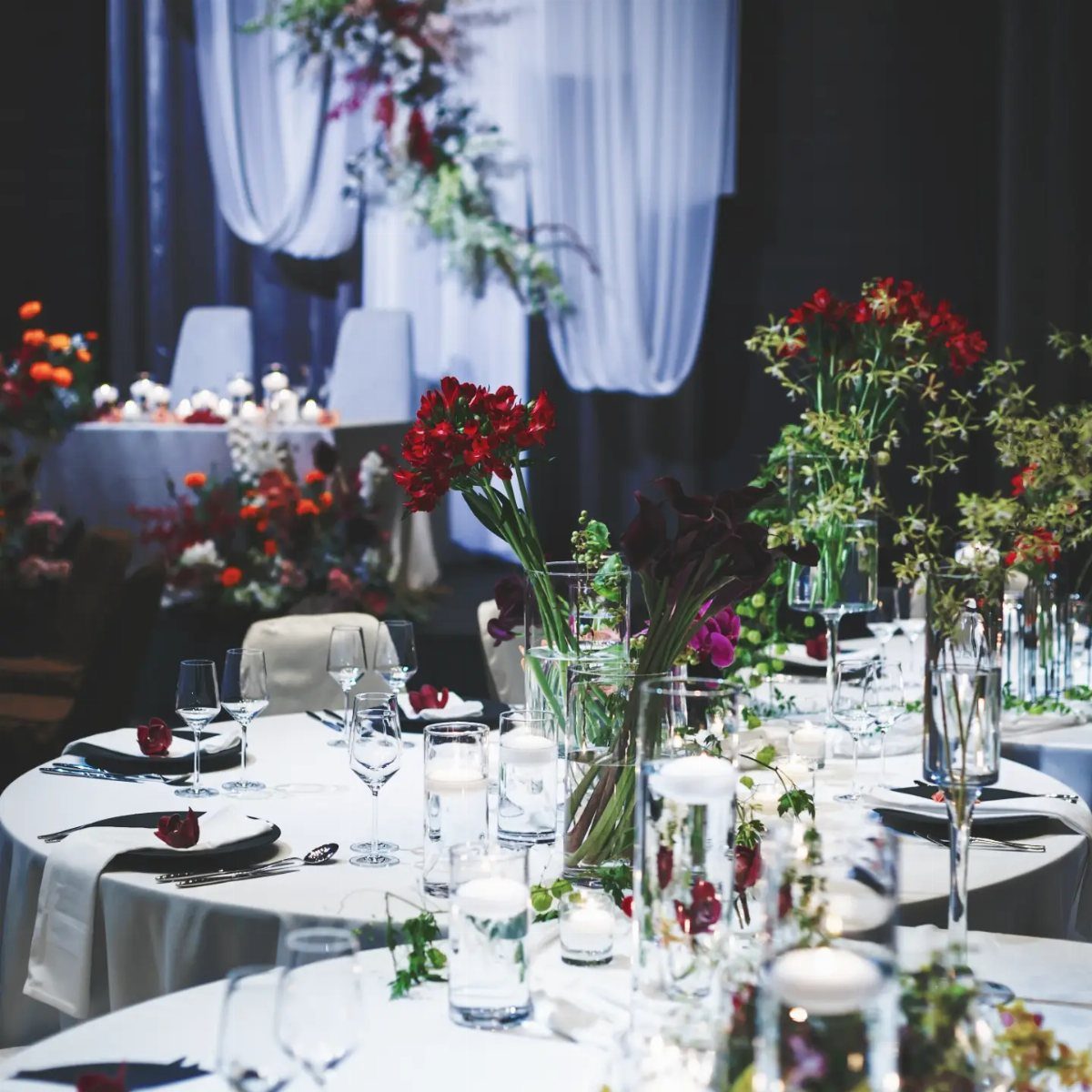 Elegant banquet tables decorated with red floral centerpieces, white candles, and glassware in a dimly lit room.
