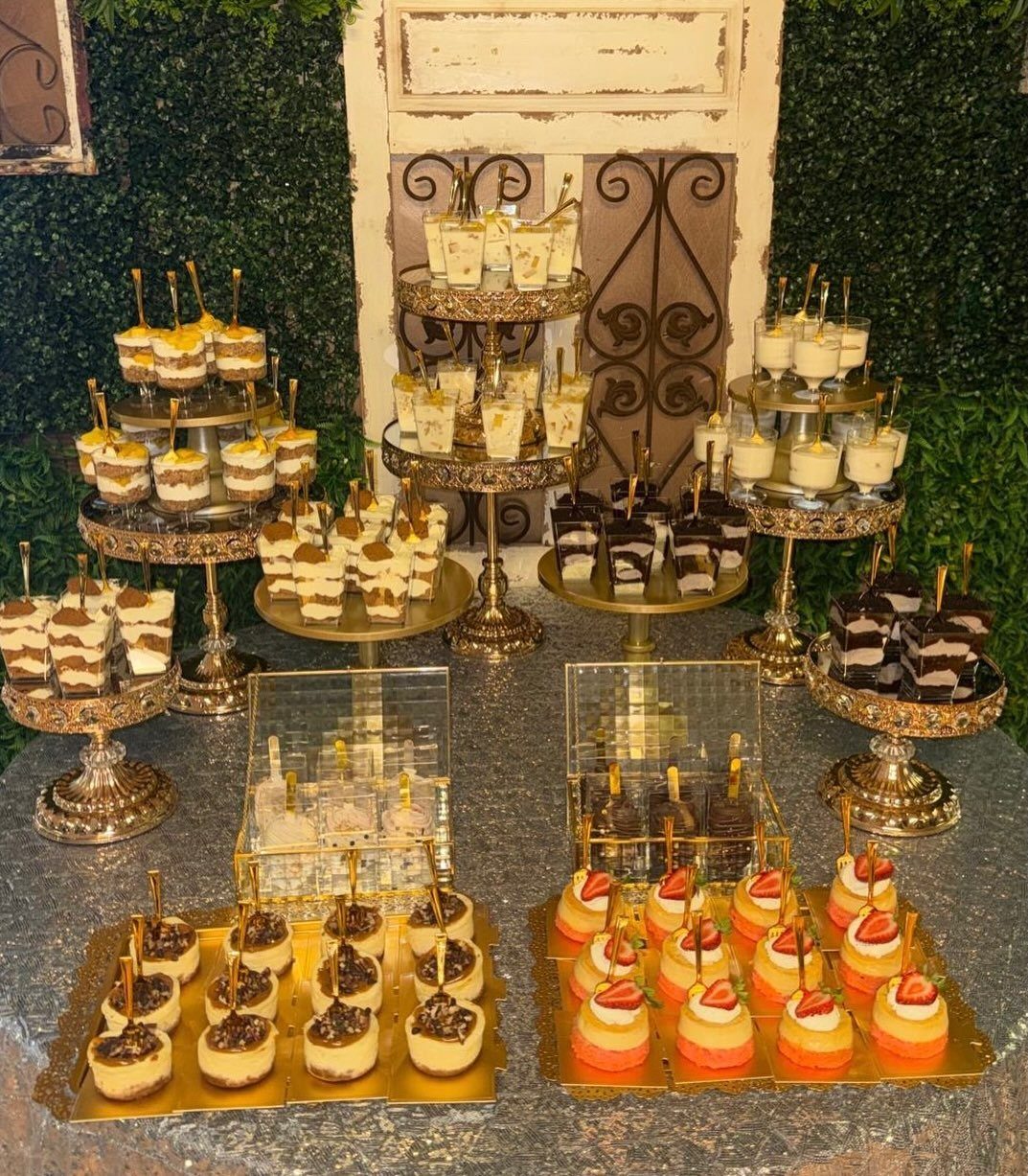 Assorted mini desserts on gold tiered stands and platters against a green hedge background.