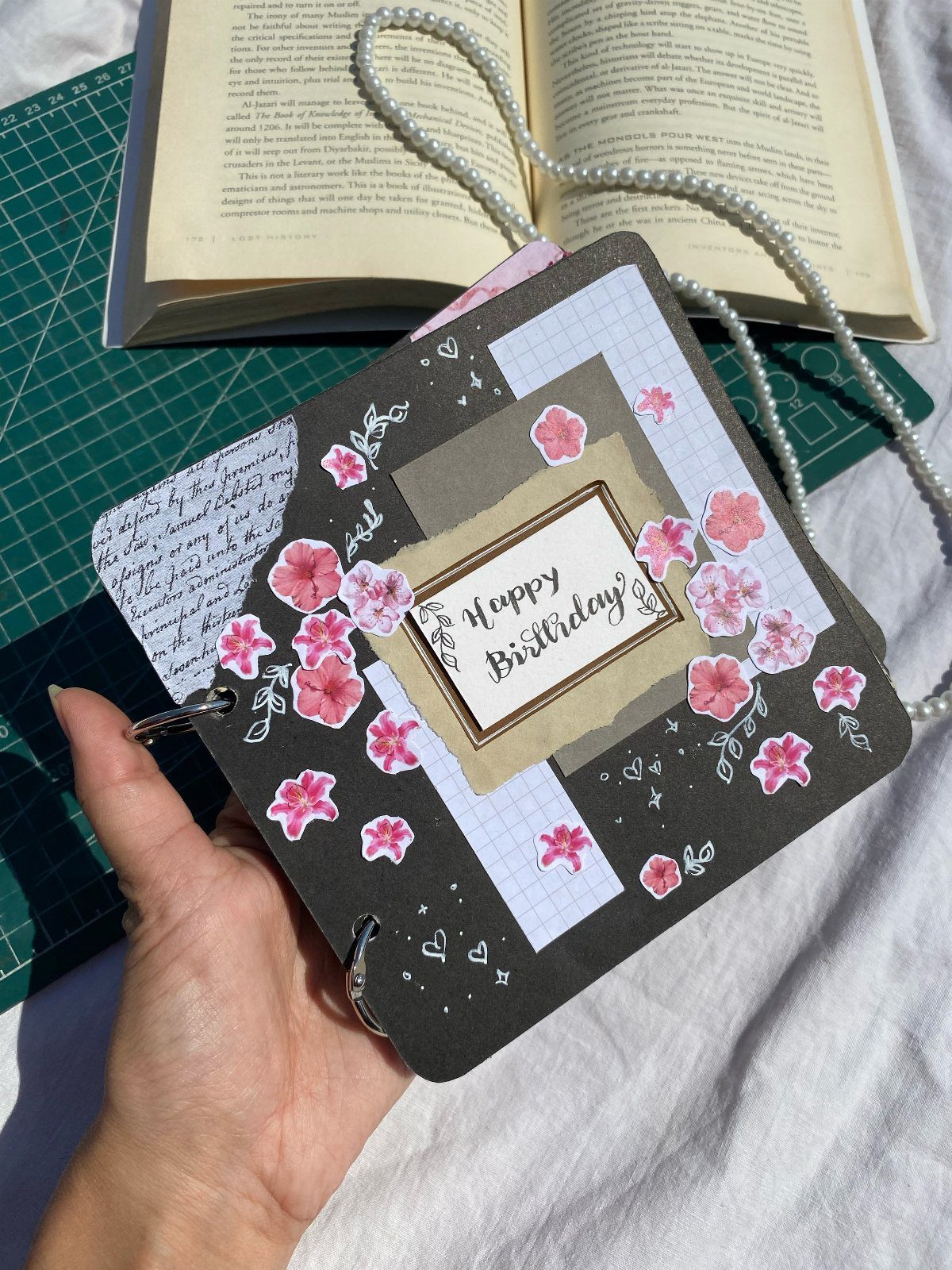A hand holding a decorated dark grey birthday scrapbook with pink floral accents.