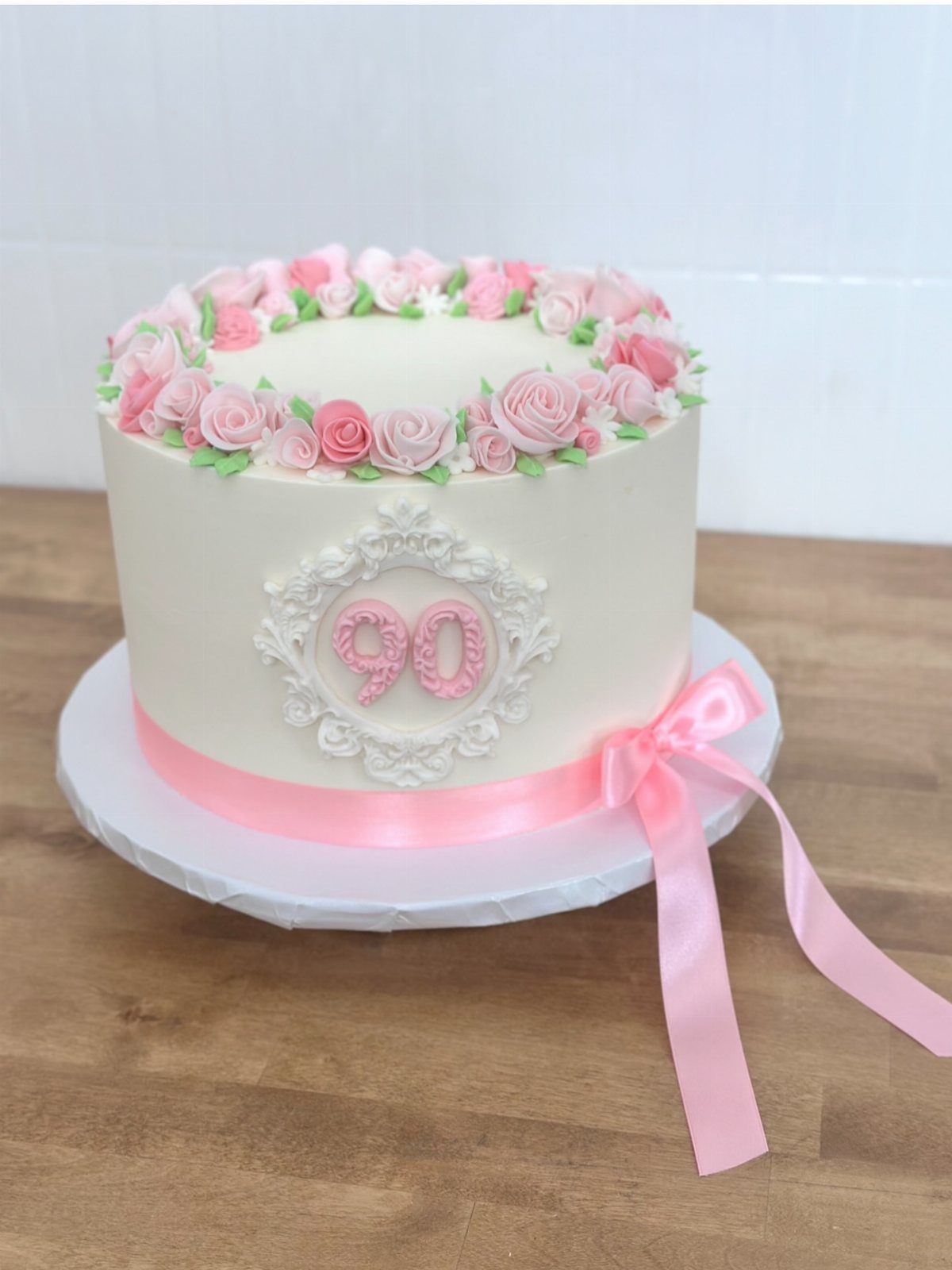 A white birthday cake decorated with pink roses and the number ninety in an ornate frame.