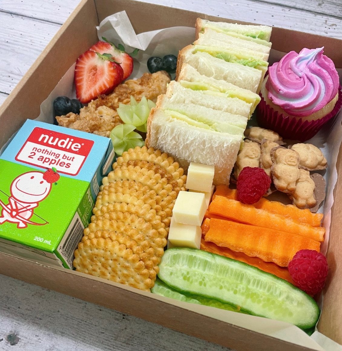 A cardboard box filled with sandwiches, crackers, vegetables, fruit, and a pink cupcake.