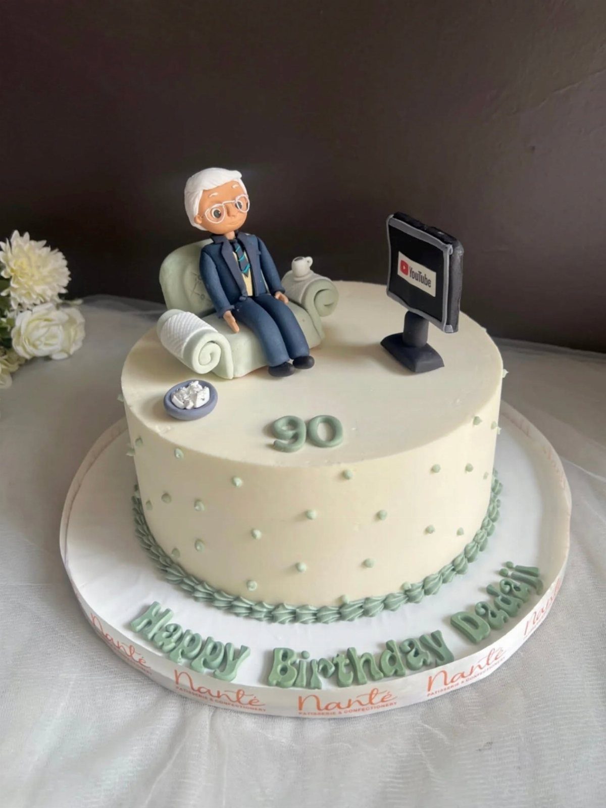 A white birthday cake featuring a figurine of an elderly man and a YouTube monitor.