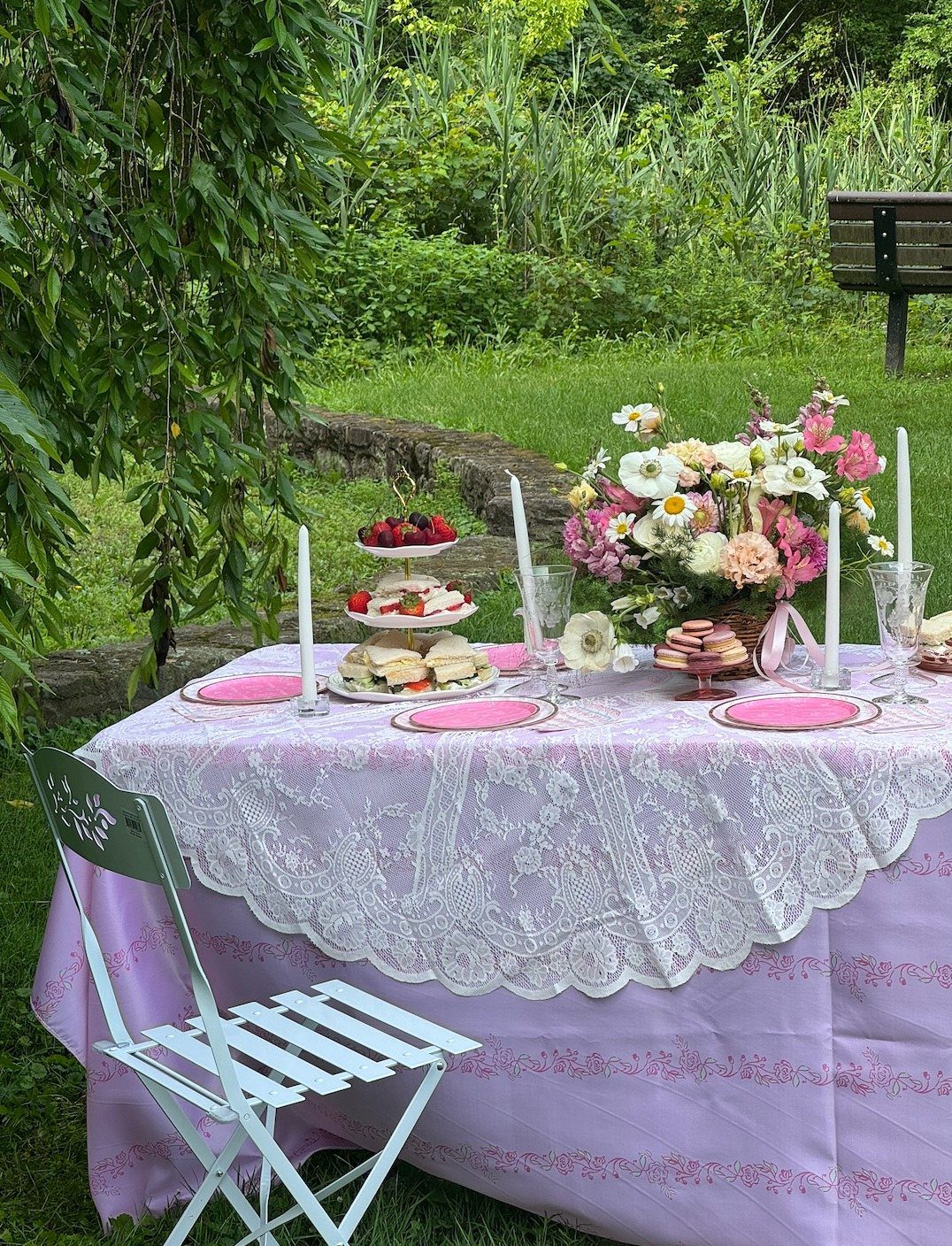 A lavender lace-covered table set with tea treats, pink plates, and flowers in a garden.