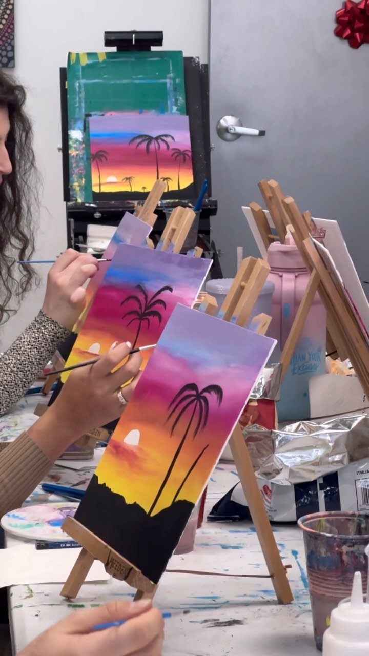 Hands of people painting sunset landscapes with palm trees on small canvases on wooden easels.