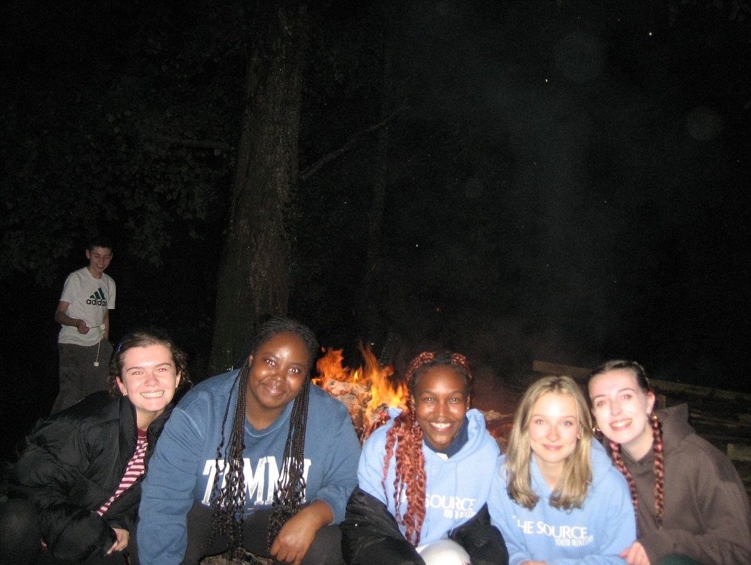 Five young women smiling together in front of a bright outdoor bonfire at night.