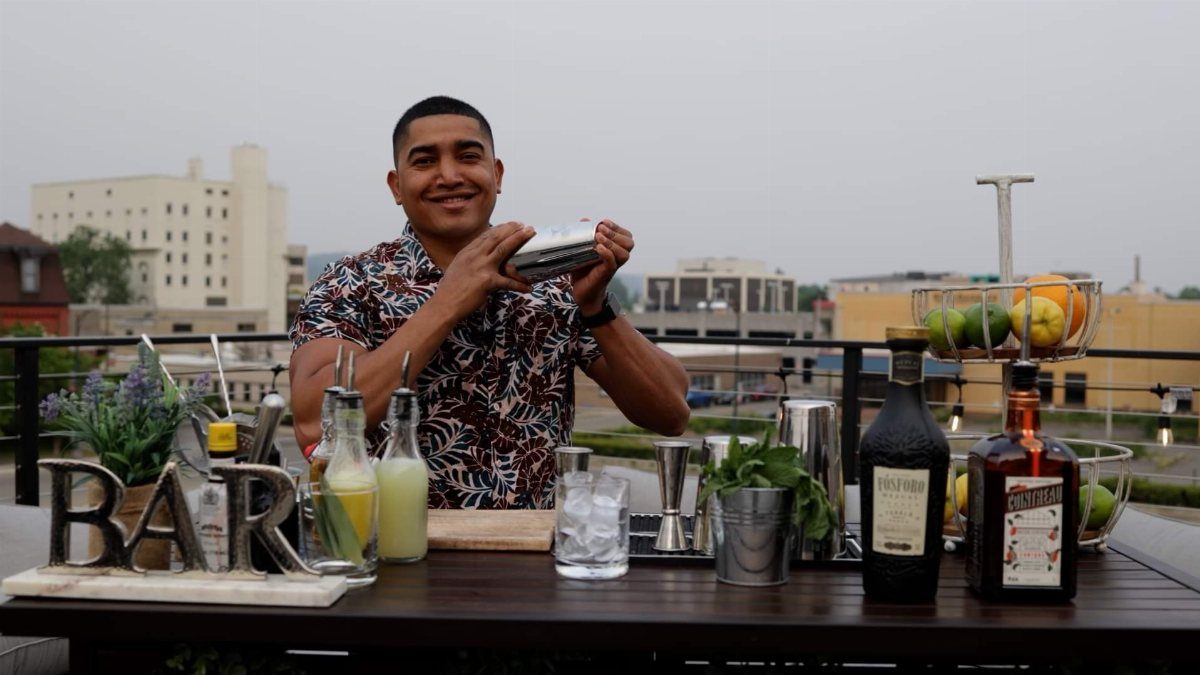A smiling man holding a cocktail shaker on a rooftop bar with various drink ingredients.