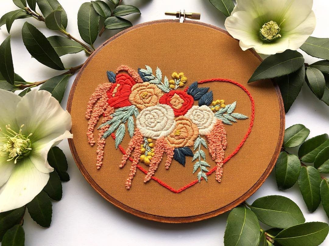 An embroidery hoop with colorful floral stitching surrounded by white flowers and green leaves.