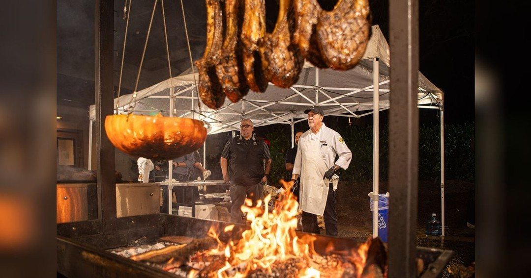 A chef stands by a large open fire pit with hanging meat.