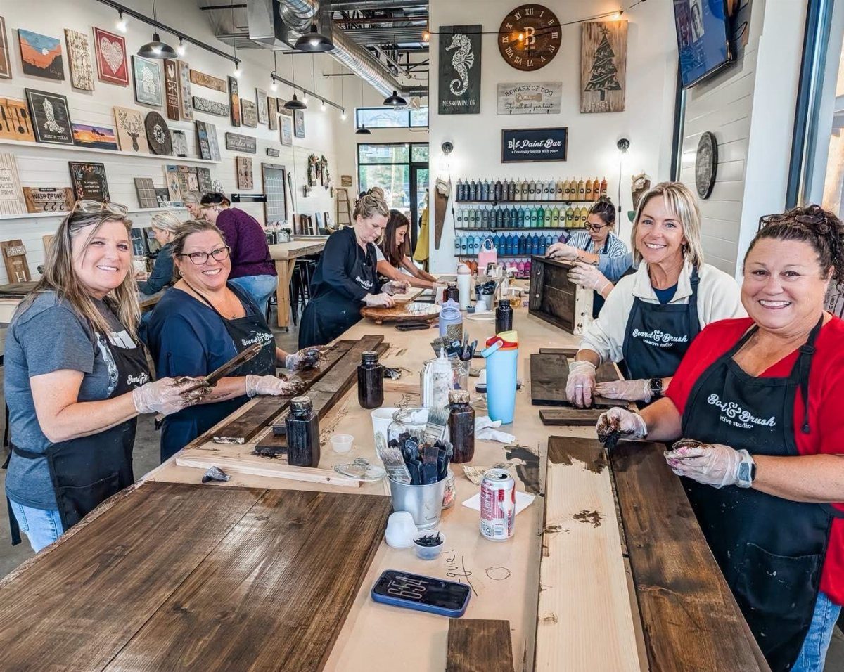 Five women in aprons smiling while working on wood crafts at a long table.