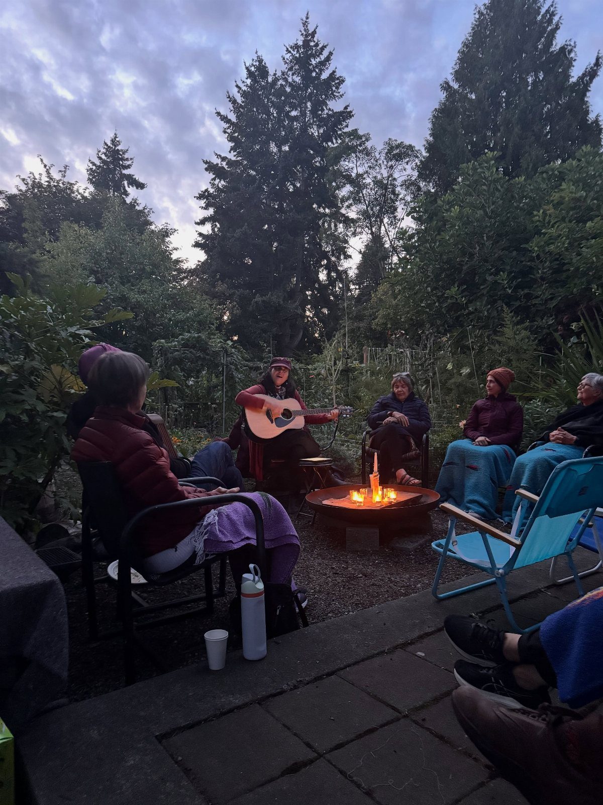 A group of people sitting around a fire pit while one person plays guitar.