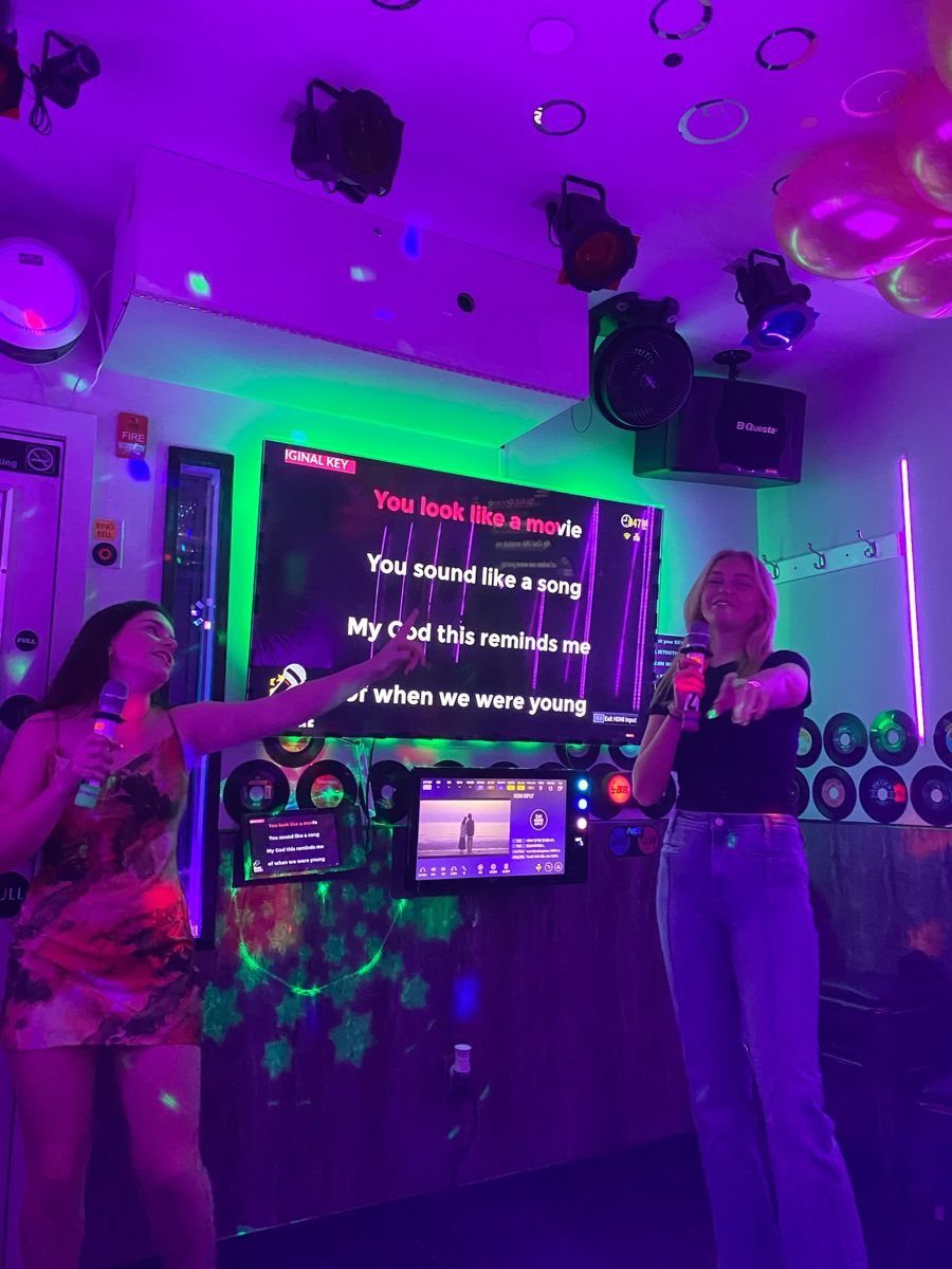 Two women singing into microphones in front of a karaoke lyric screen with neon lighting.
