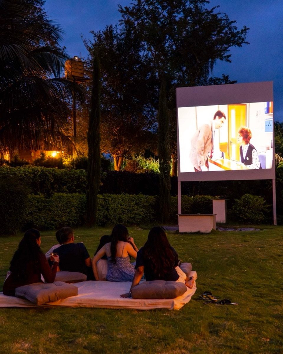 Four people sitting on a blanket on a lawn watching an outdoor movie screen at night.