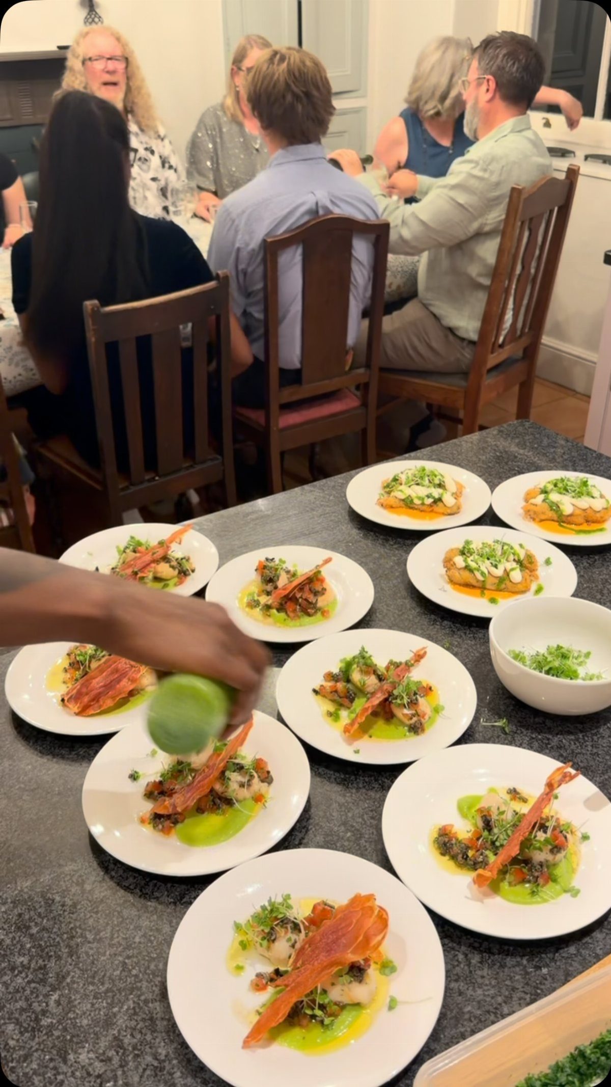 A chef garnishing multiple plates of seafood appetizers on a dark kitchen counter.