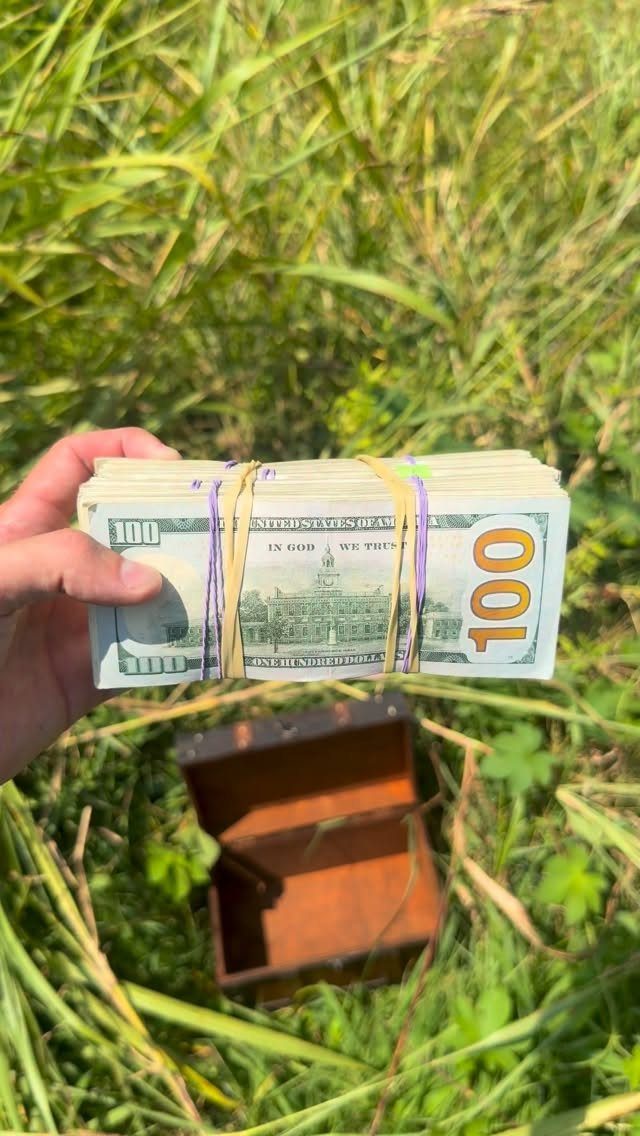 A hand holding a stack of hundred dollar bills above an open wooden treasure chest in grass.