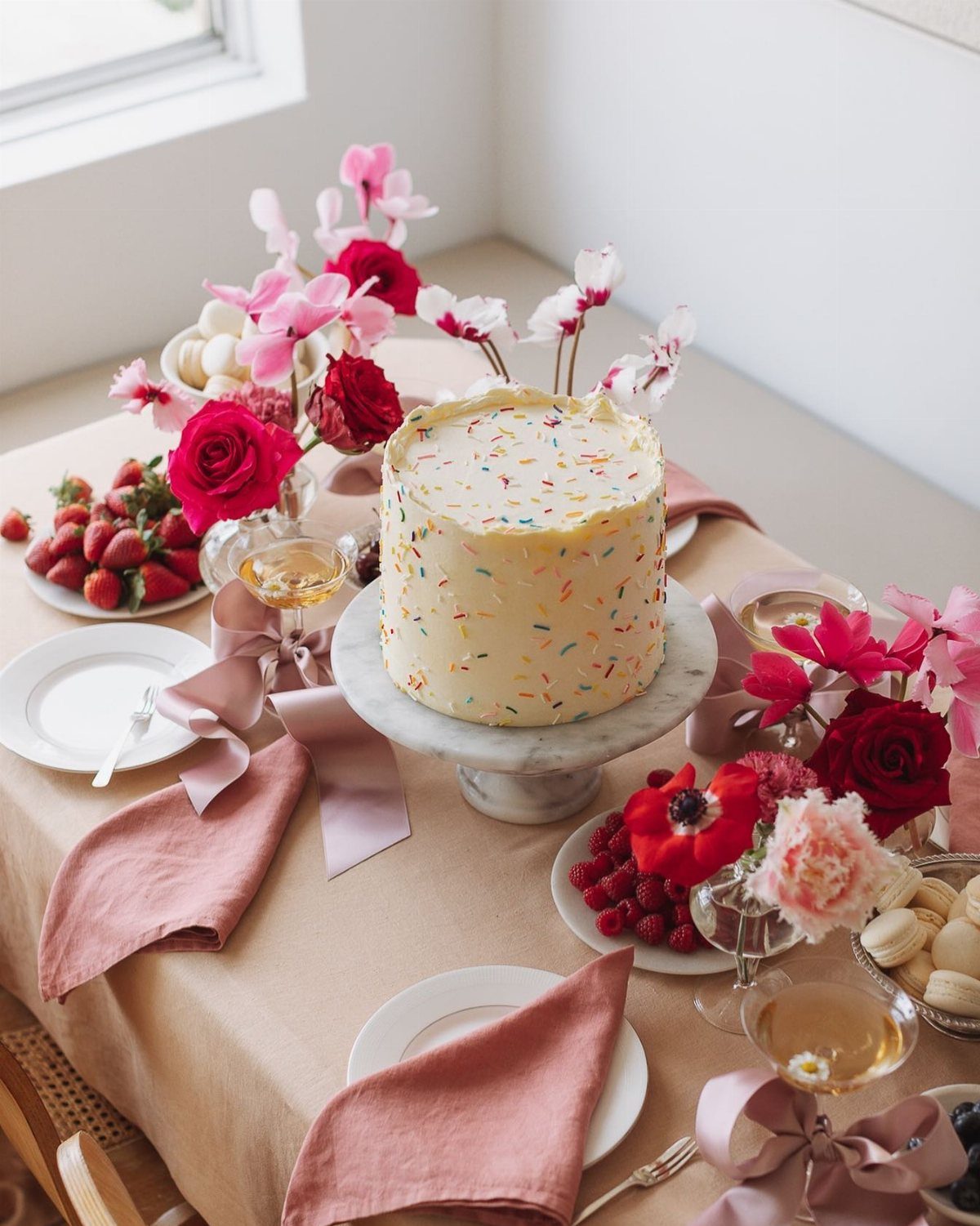 A sprinkle-covered white cake on a marble stand surrounded by roses and fruit.