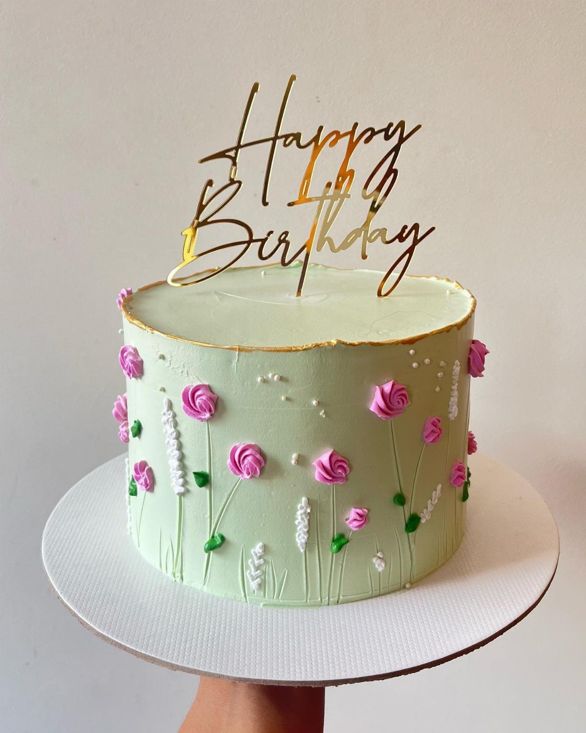 Light green cake decorated with 3D pink frosting flowers and a gold Happy Birthday topper.