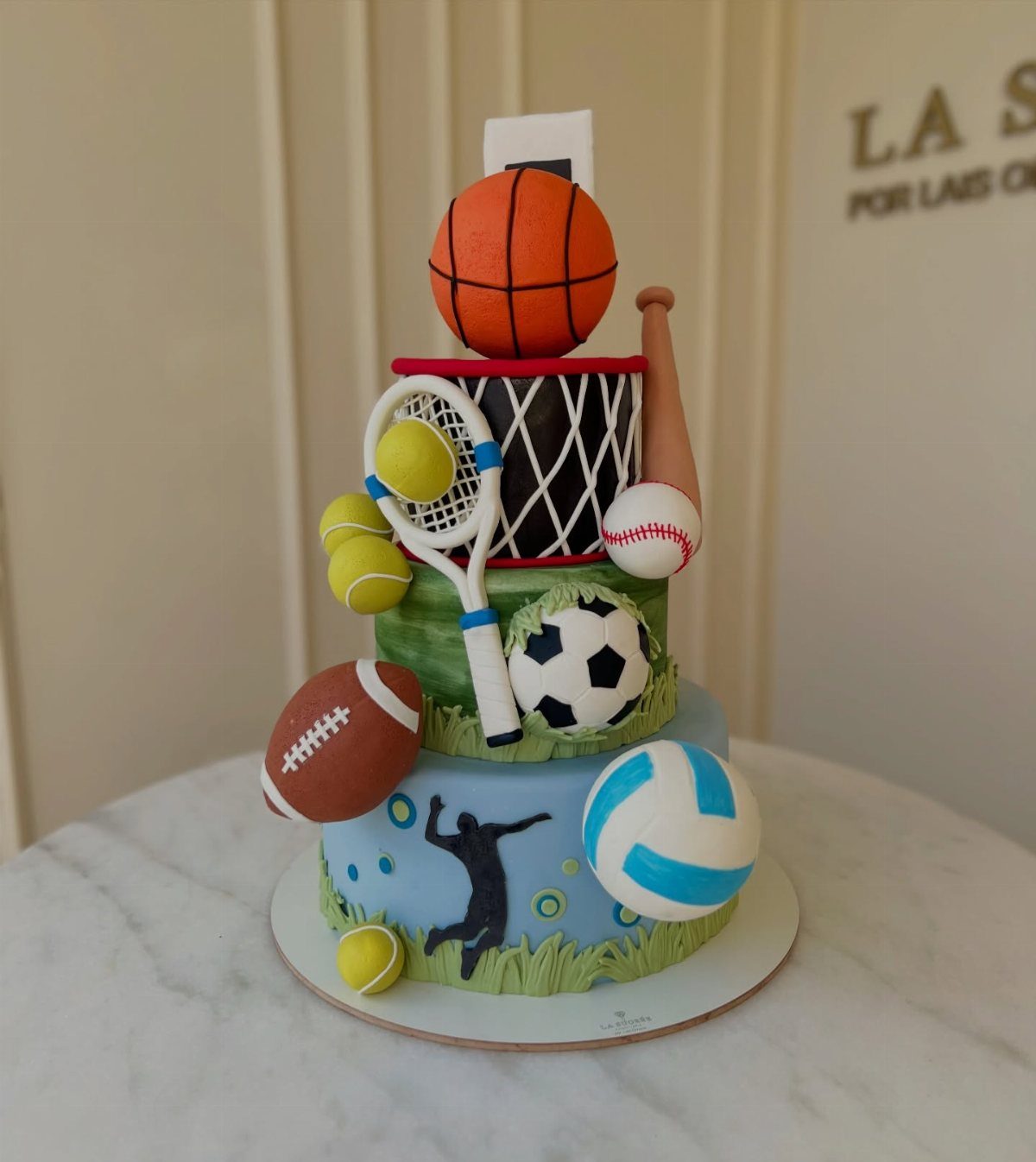 A multi-tiered cake decorated with various edible sports balls and equipment like basketballs and tennis rackets.