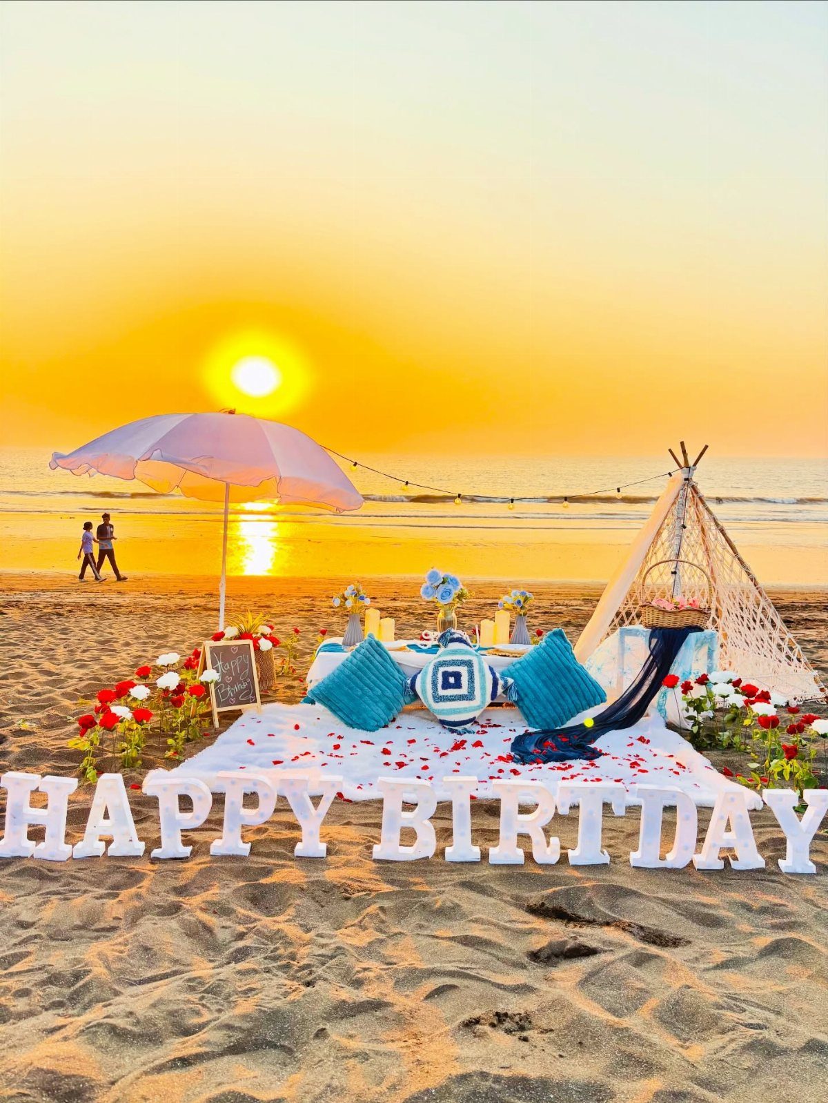 Beach picnic setup with pillows, a teepee, and white birthday letters at sunset.