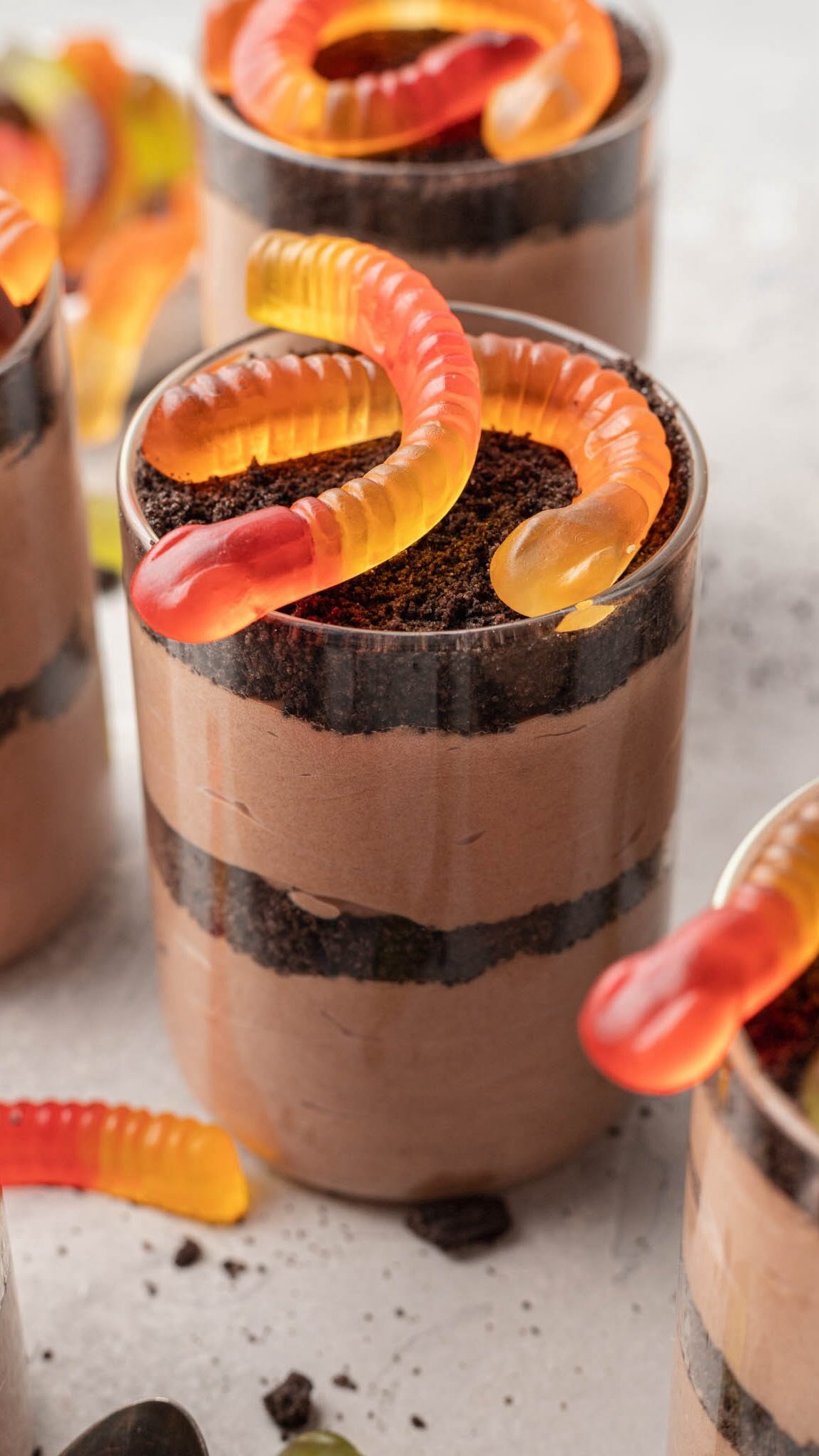 Layered chocolate mousse and cookie crumb desserts topped with colorful gummy worms in clear glasses.