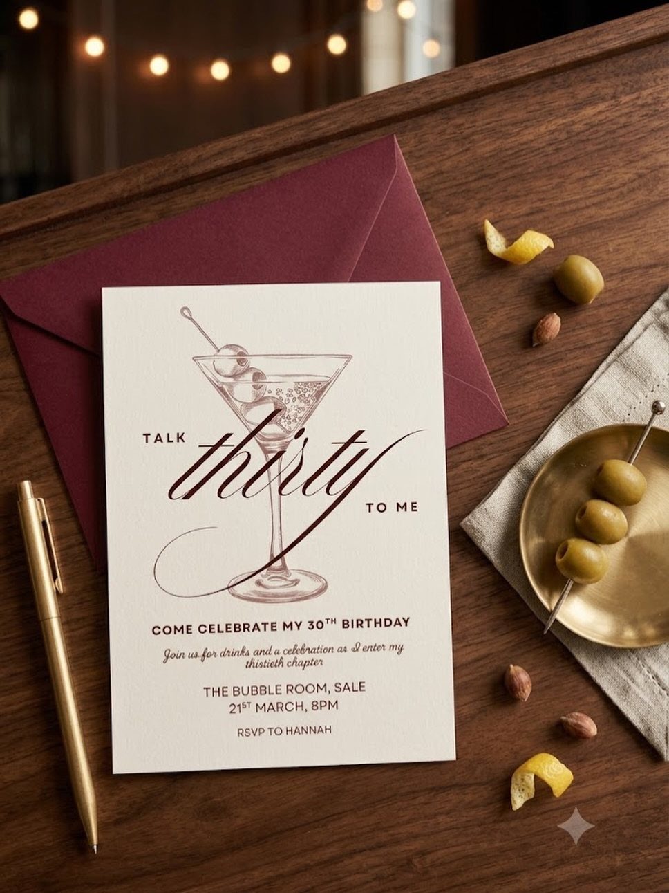 A white martini themed birthday invitation card resting on a wooden table with olives and a pen.