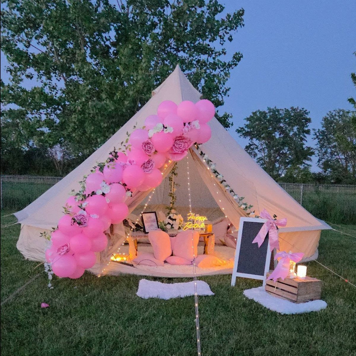 A cream bell tent decorated with pink balloons and pillows on a grassy lawn.