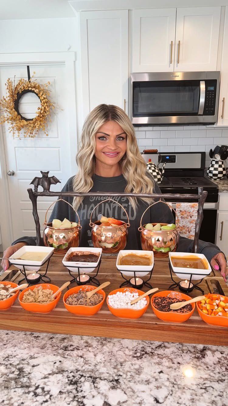A woman presenting a caramel apple bar with sliced apples and various toppings in a kitchen.